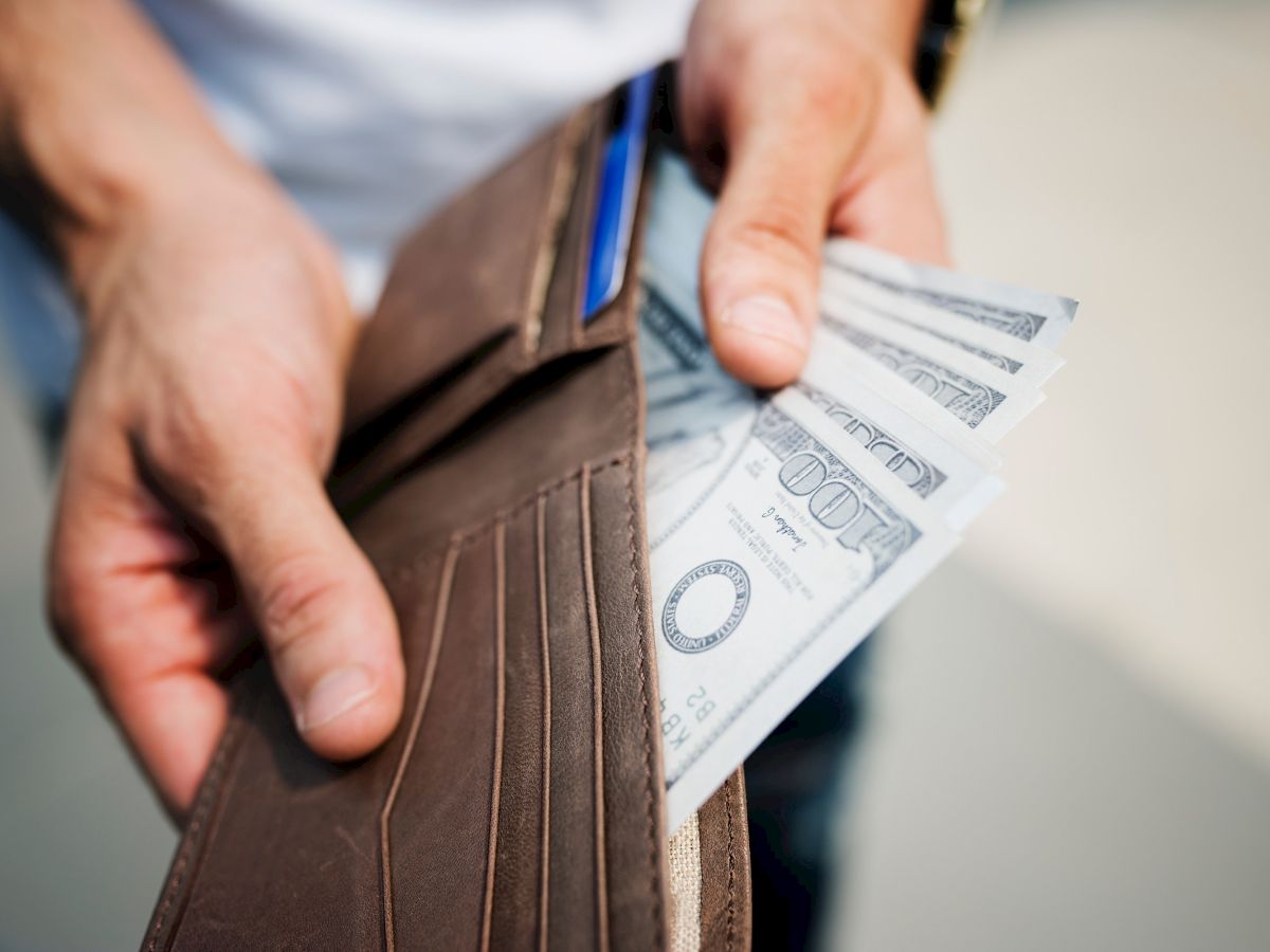 A person is holding a brown wallet in their hands, with several 100-dollar bills partially visible inside the wallet.