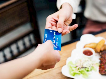 Two people are exchanging a blue card over a table with food, including salad, grilled items, and a sauce bowl near the edge of the table.