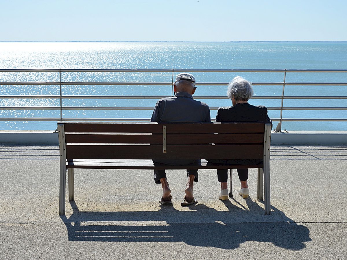 Two people sit on a bench facing a calm sea, basking in the peaceful scenery on a sunny day.