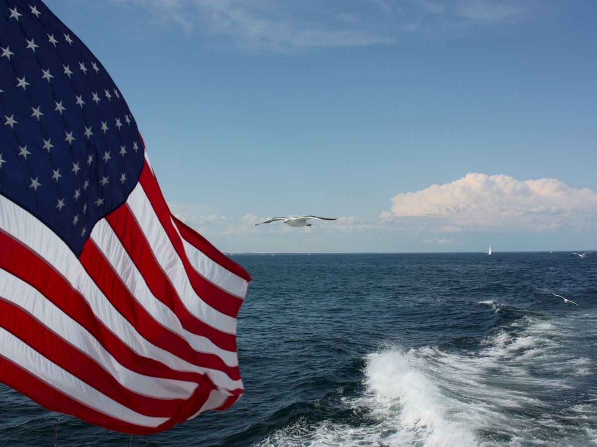 An American flag flutters while a seagull flies over the ocean, leaving a boat's wake against a backdrop of clear skies and distant clouds.