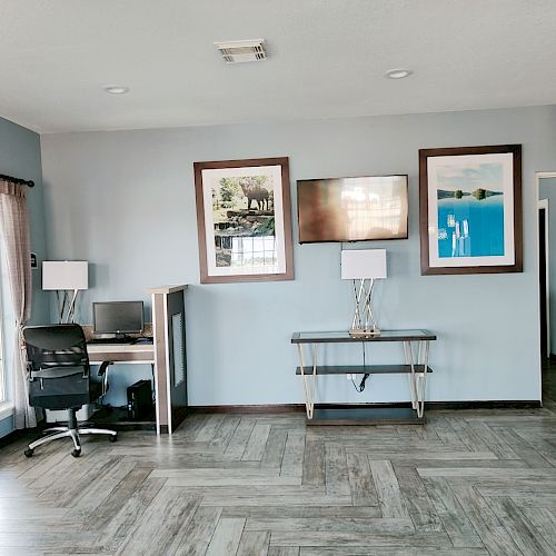 The image shows a clean, modern room with a desk, office chair, two lamps, and framed pictures on the wall. There's a large window on the left.