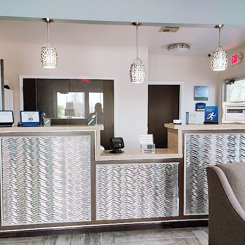 The image shows a reception desk area with modern design elements, including pendant lights, a patterned panel, and an adjacent waiting area with seating.