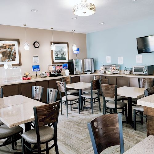 A small dining area with multiple tables and chairs, a breakfast buffet with various items, a TV on the wall, and decorative framed pictures.