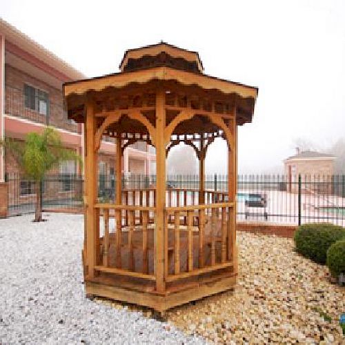 The image shows a wooden gazebo in the foreground, with a building and a fenced area with greenery in the background.