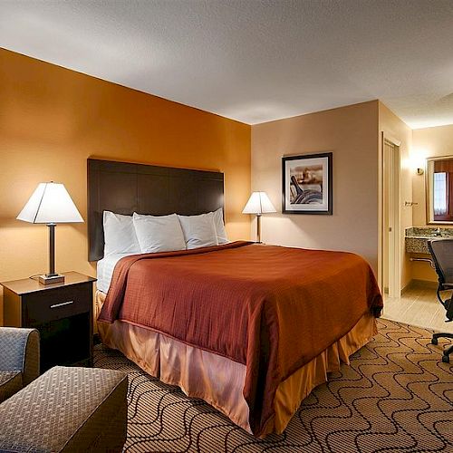 This image shows a cozy hotel room with an orange accent wall, a bed, lamps, a chair, a desk, TV, and a partial view of the bathroom area.