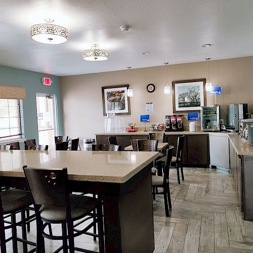 A small dining area with tables, chairs, a countertop, coffee machines, and a television on the wall. The room is well-lit with a modern design.