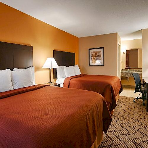 This image shows a hotel room with two double beds, orange bedding, nightstands with lamps, a TV on a dresser, and a desk with a lamp in the back.