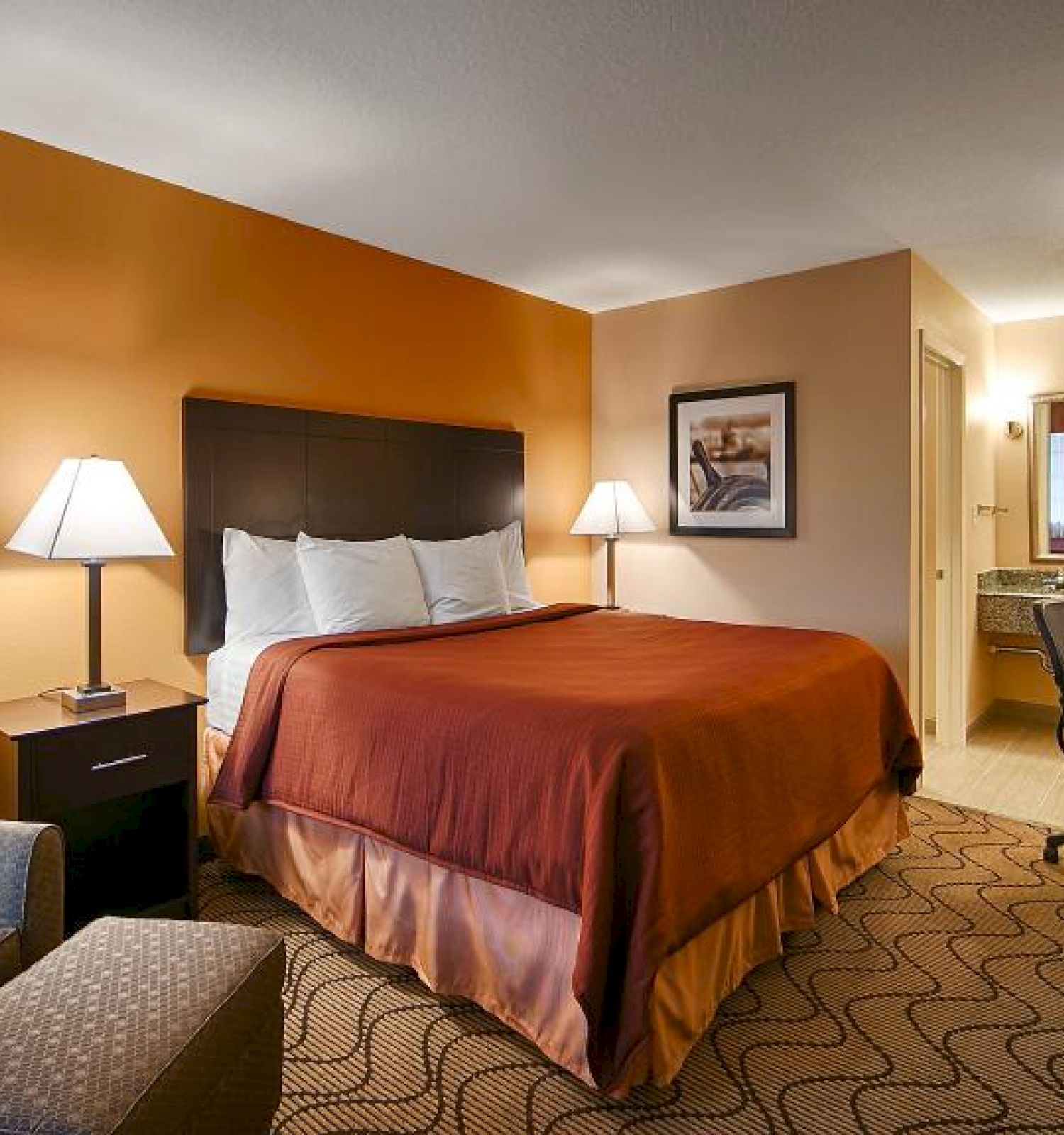 This image shows a well-lit hotel room with a double bed, armchair, nightstands, lamps, a desk chair, a large TV, and an adjacent bathroom.