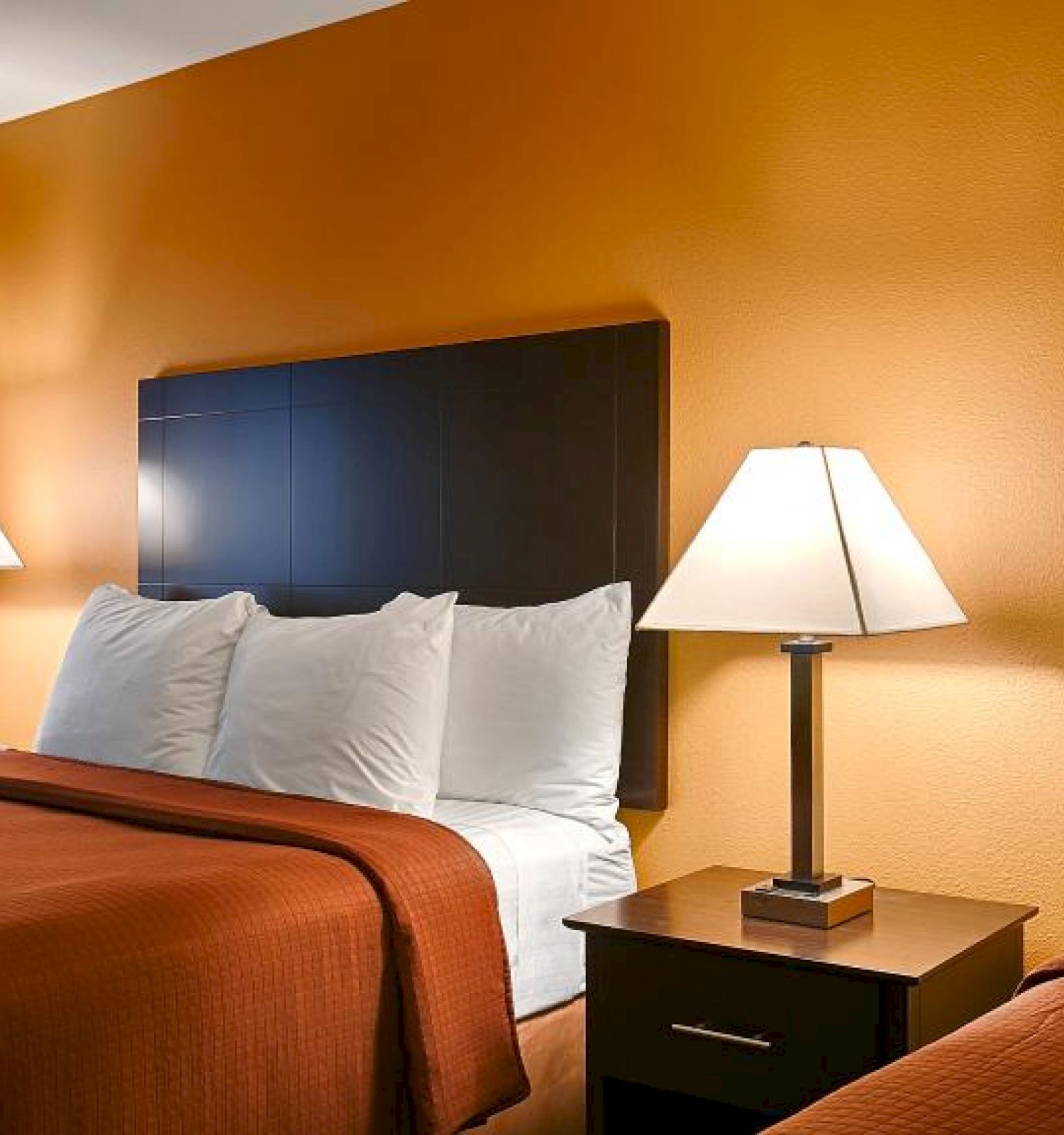 This image shows a hotel room with two beds, each having white pillows and brown covers, two lamps on nightstands, and orange walls.