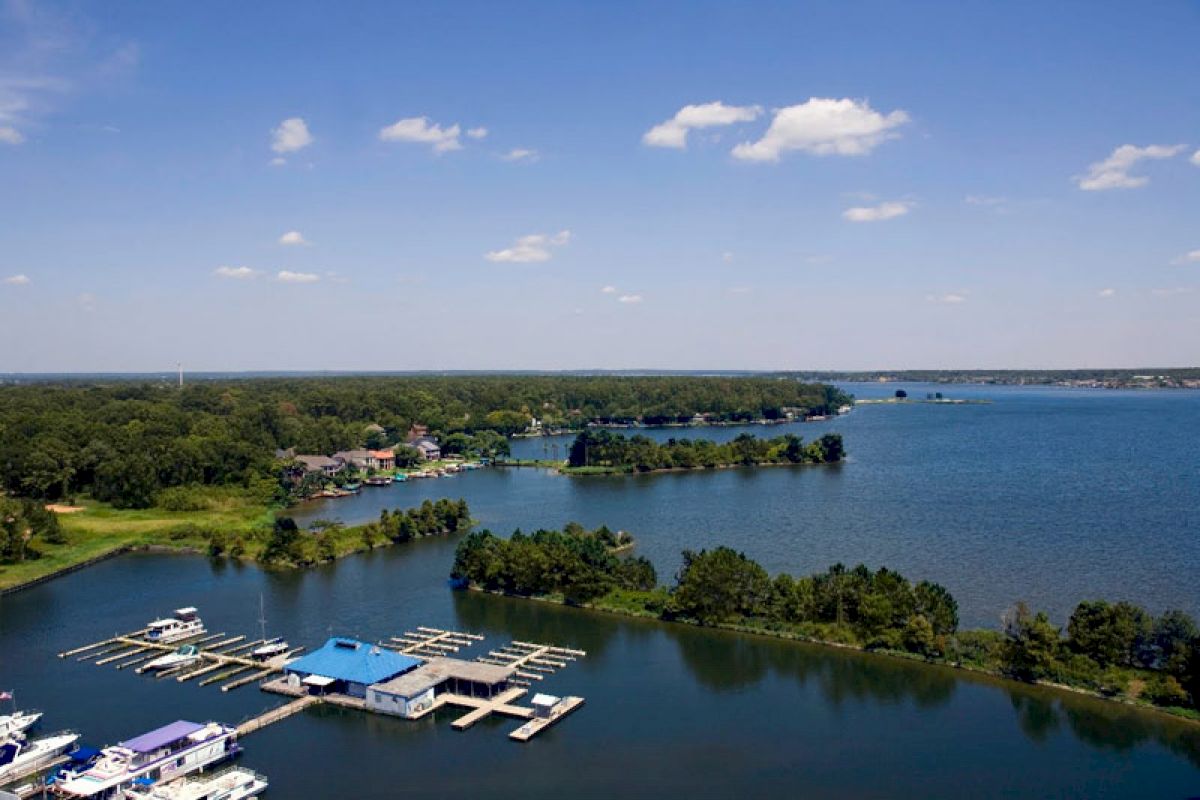 An aerial view of a marina on a lake with docks, boats, surrounding greenery, and a distant horizon under a blue sky with scattered clouds.