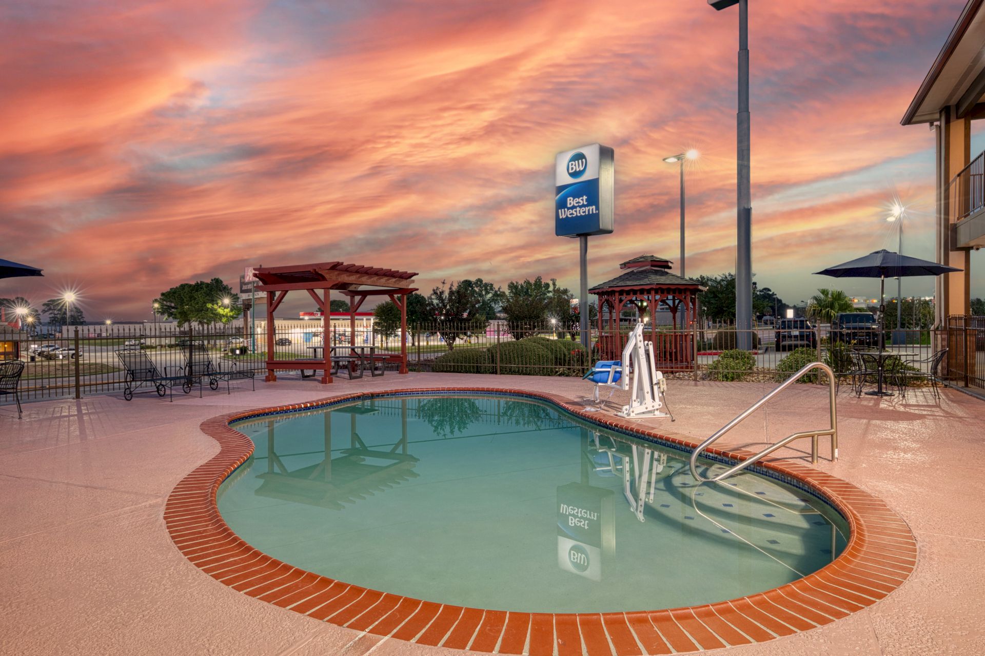 An outdoor hotel pool at dusk with a &ldquo;Best Western&rdquo; sign, umbrellas, loungers, and trees under colorful sunset clouds.