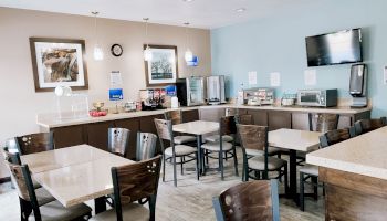 A small dining area with tables, chairs, a TV, coffee station, and breakfast food setup. Paintings and lights decorate the room.