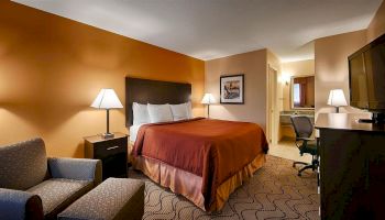 A cozy hotel room with an orange accent wall, a queen-sized bed, lamps, an armchair, a desk, a wall-mounted TV, and an open bathroom door.