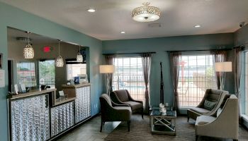 A modern waiting area with a reception desk, patterned counter, seating, lamps, rug, and large windows with drapes, under a stylish ceiling light.