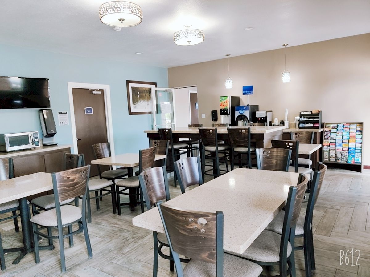 The image shows a small dining area with multiple tables and chairs, a TV on the wall, a coffee station, and a breakfast bar setup.