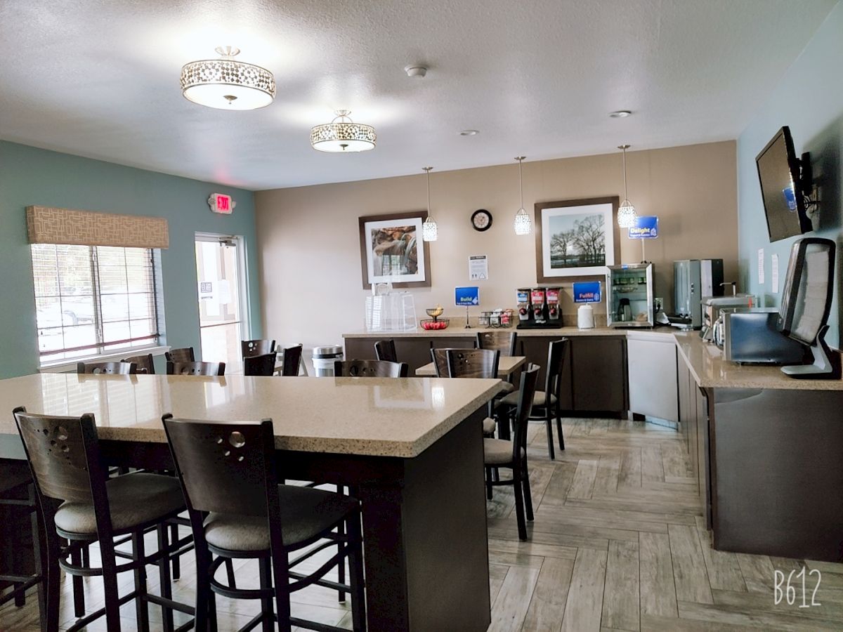 A modern dining area with tables, chairs, a TV, coffee station, and artwork on the walls. Brightly lit with windows and ceiling lights.