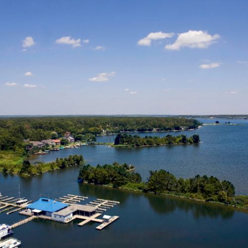 The image shows a scenic view of a lake with docks, boats, a blue-roofed building, and lush green landscape under a partly cloudy sky.