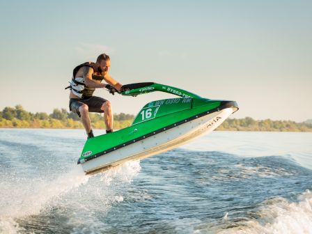 A person riding a green jet ski numbered 16, performing a jump over water with a life vest on, set against a clear blue sky and distant foliage.