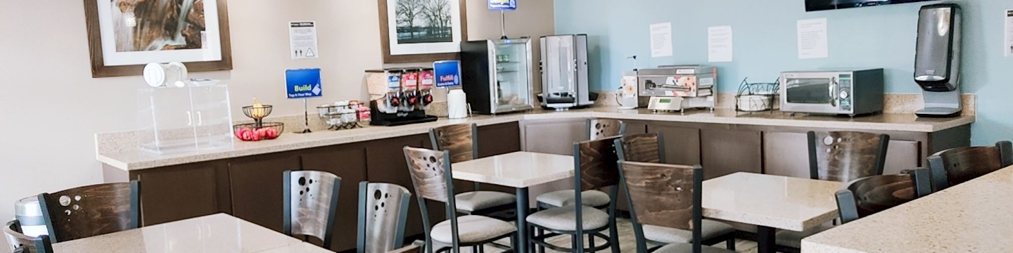 The image shows a dining area with various tables and chairs, a breakfast counter with food and beverage options, and a wall-mounted TV.