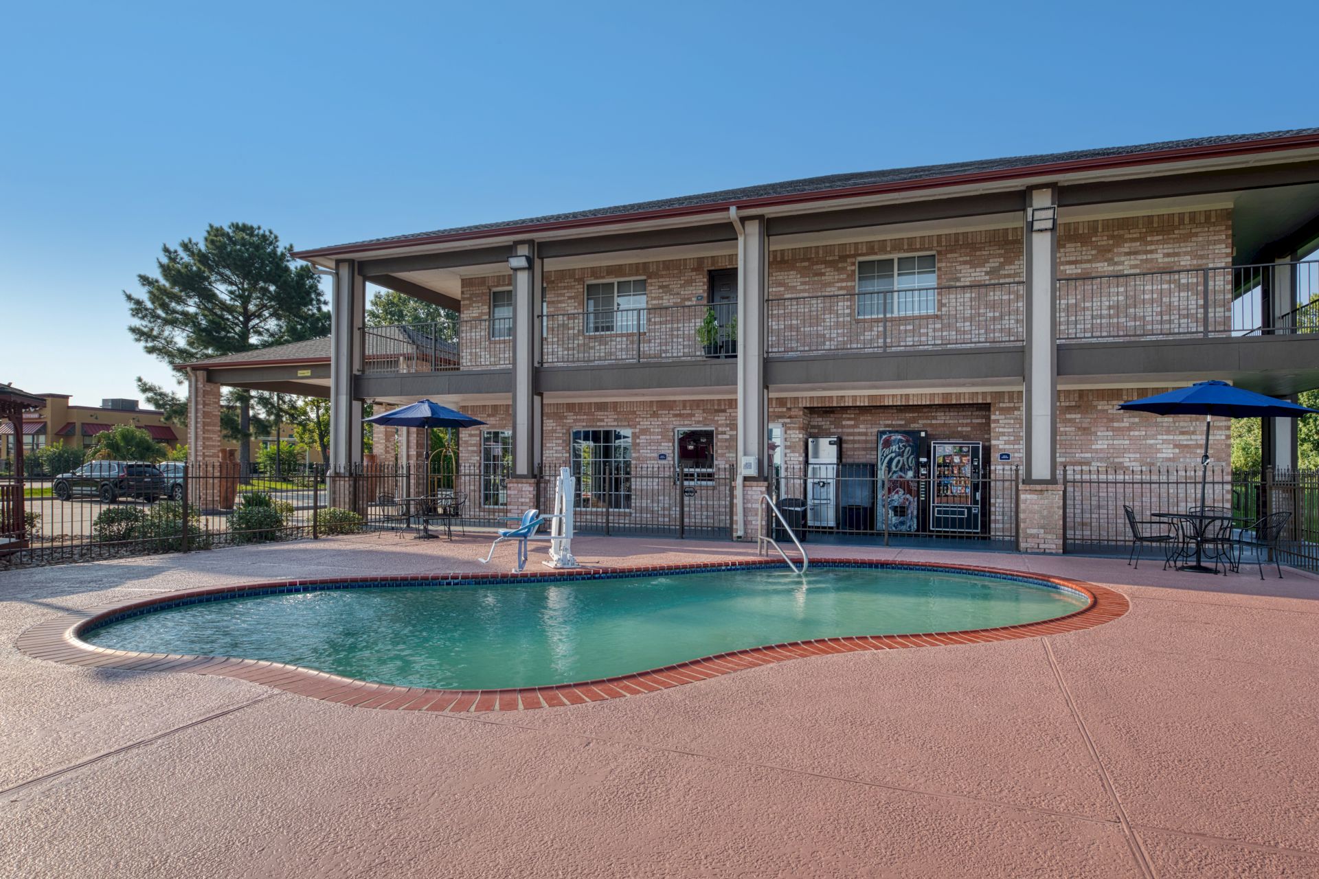A sunny outdoor swimming pool in front of a brick apartment complex with chairs, umbrellas, and a fenced patio area.
