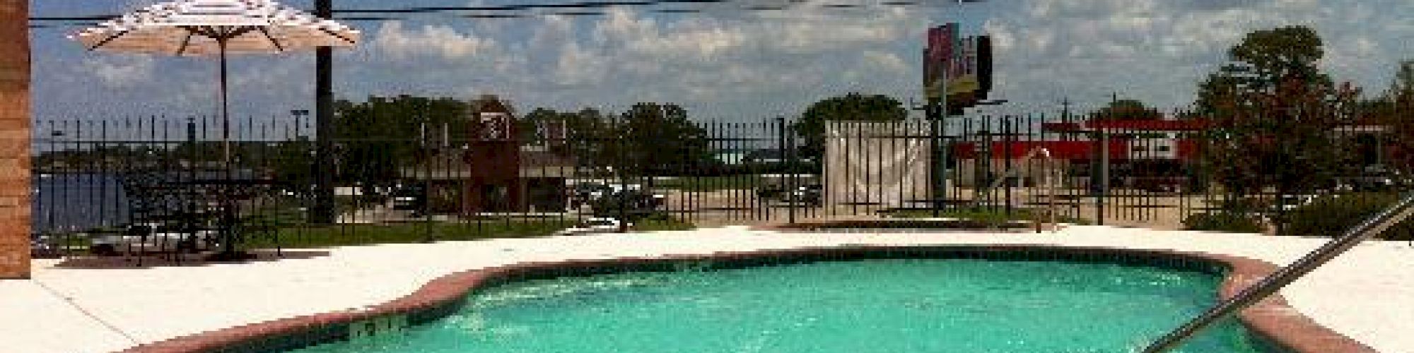 A small, outdoor swimming pool with a fence, umbrella, and partially cloudy sky in the background. A red logo sign is visible in the distance.