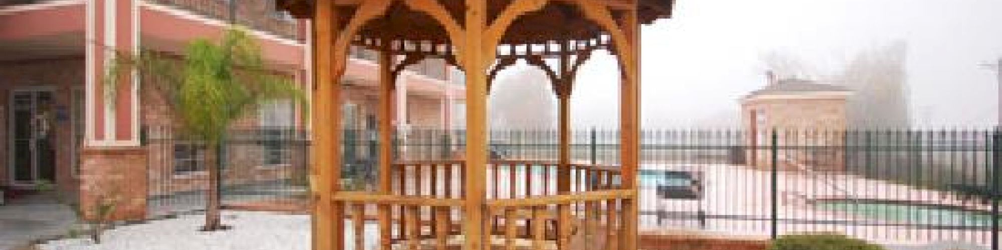 The image shows a wooden gazebo in a landscaped area with white gravel, near a building and a fenced area with landscaping greenery.